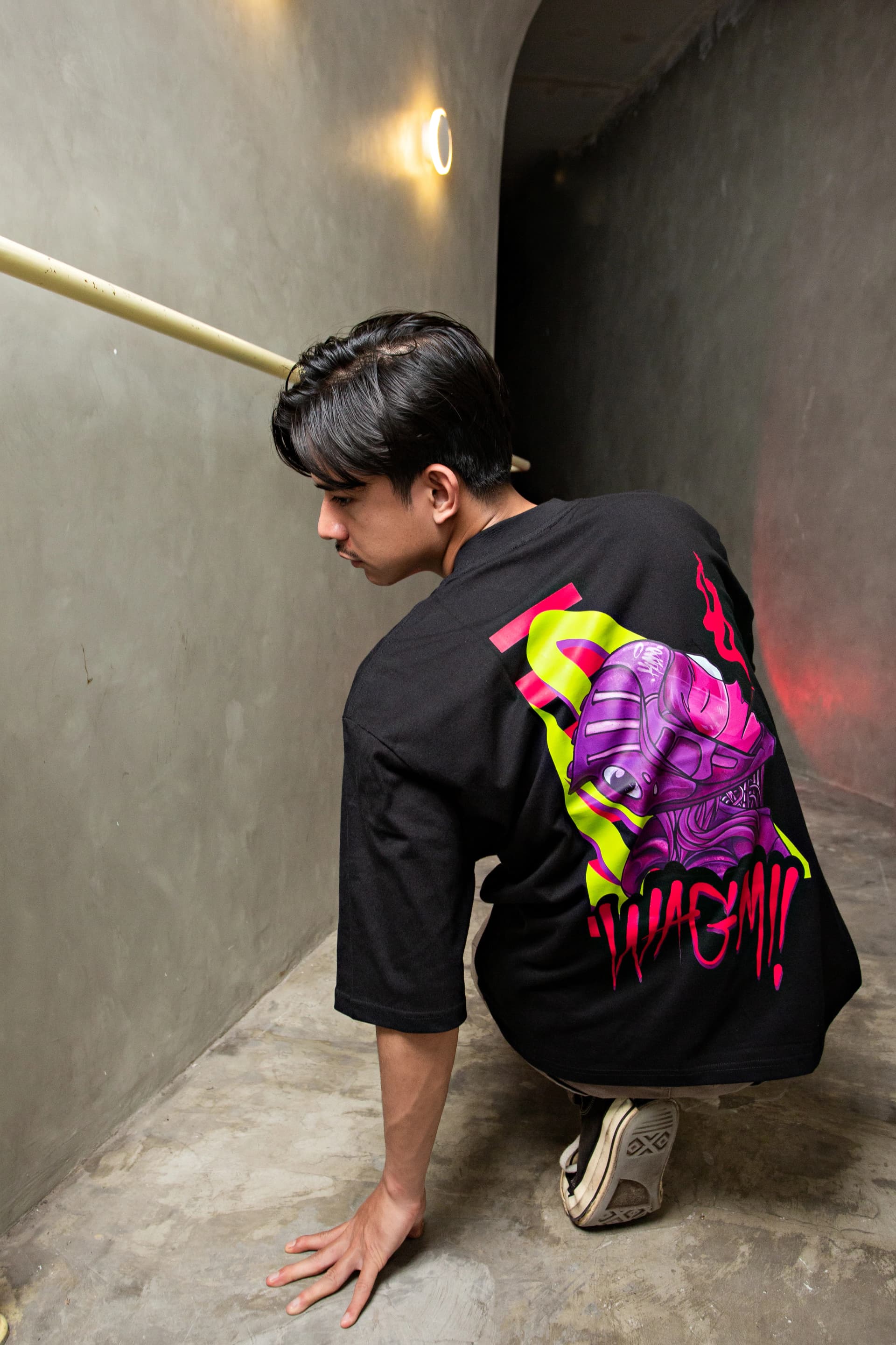 Man crouching in a concrete corridor wearing a black T-shirt with a colorful back graphic