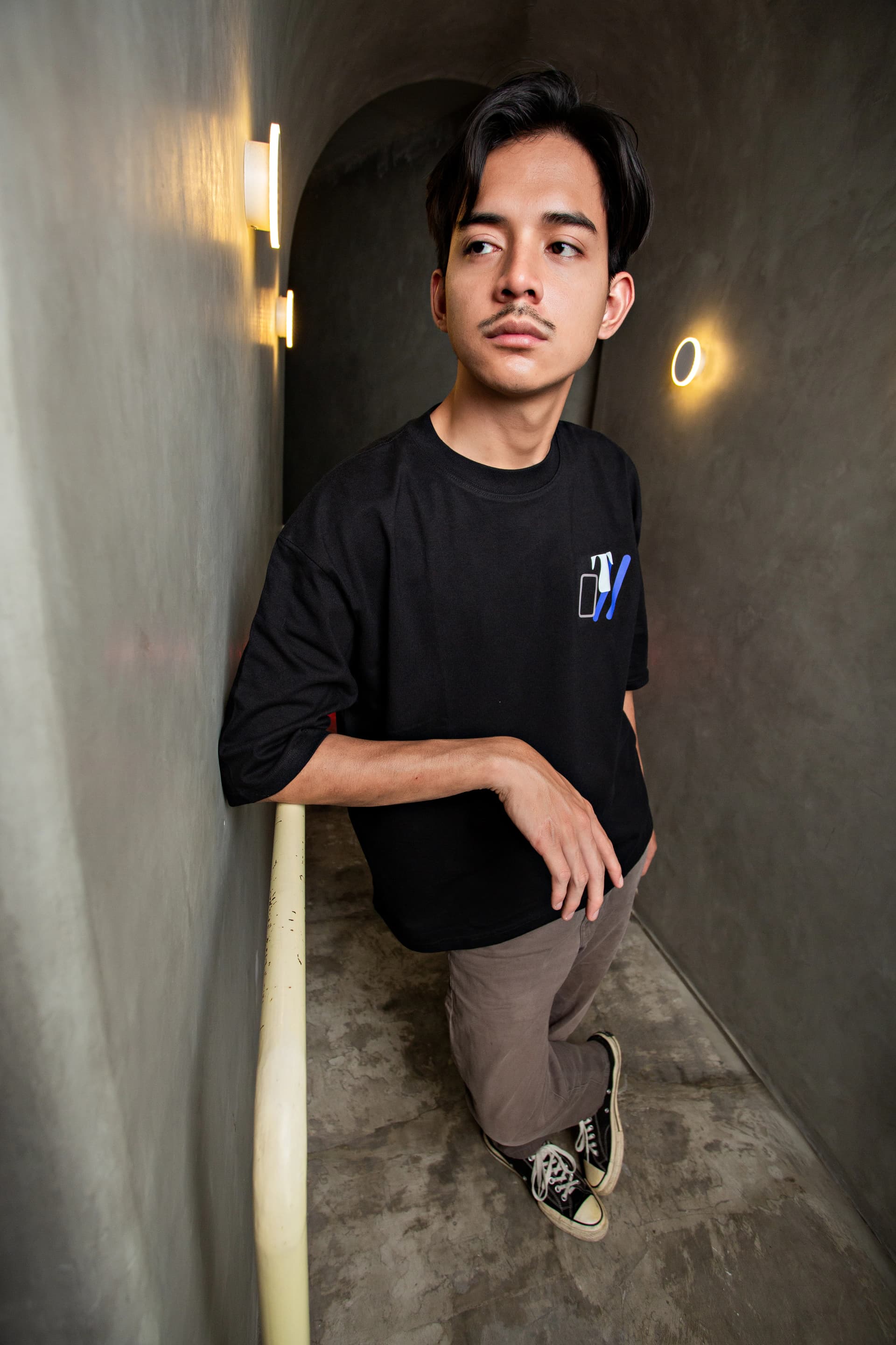 Man leaning on a corridor railing in a black oversized T-shirt and gray trousers