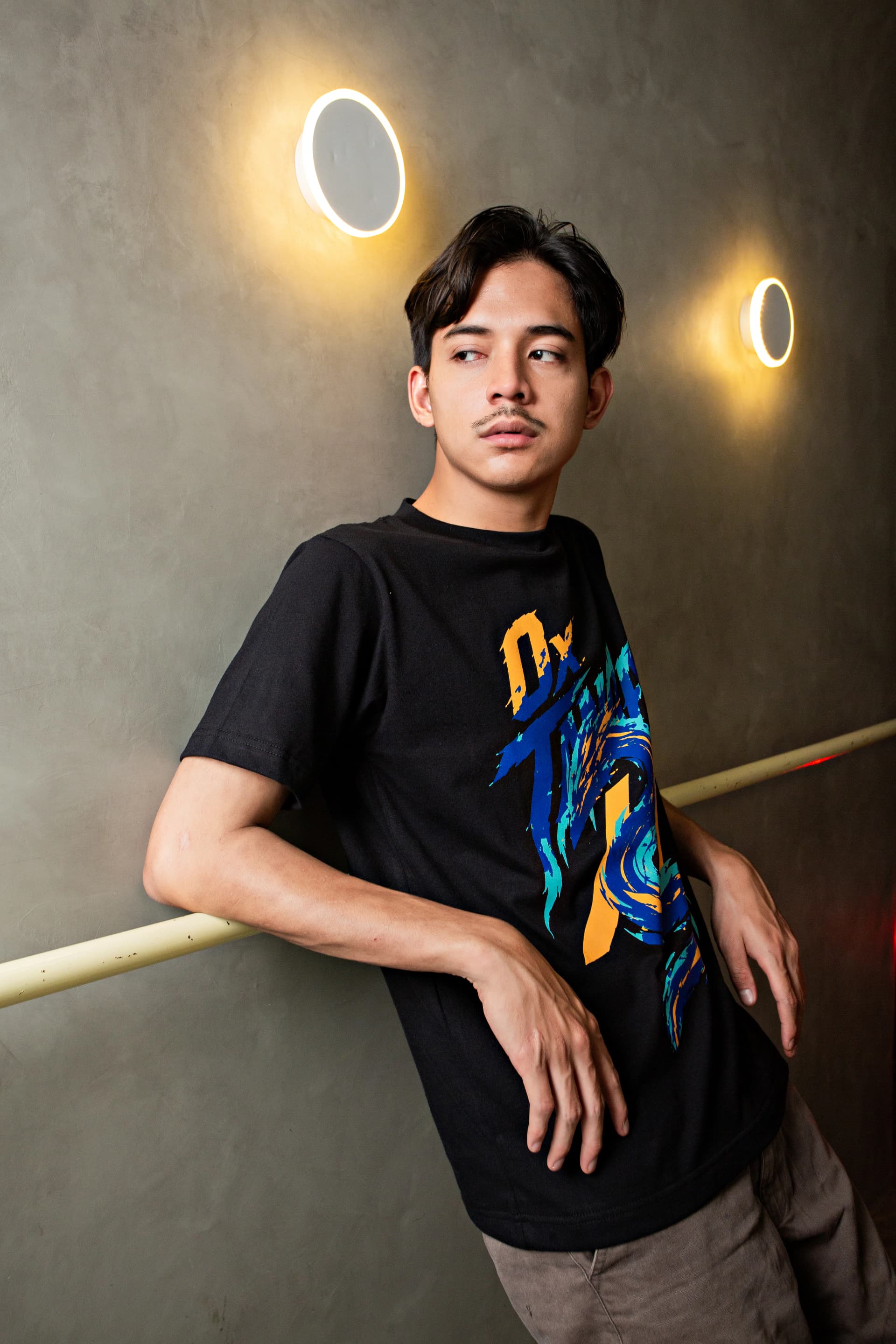 Man leaning against a railing in a black graphic T-shirt inside a concrete corridor
